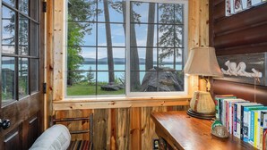 Ein Holzhütten-Innenraum mit großem Fenster mit Blick auf einen See und Bäume, einer Lampe und einem Bücherregal.