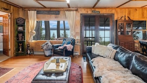 Ein gemütliches Blockhaus-Interieur mit Kamin, einer Ledersofa, einem Lesesessel und Blick auf den See.