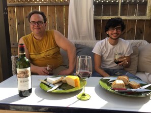 Two people seated at an outdoor table with a bottle of wine, cheese, bread, and a glass of wine.