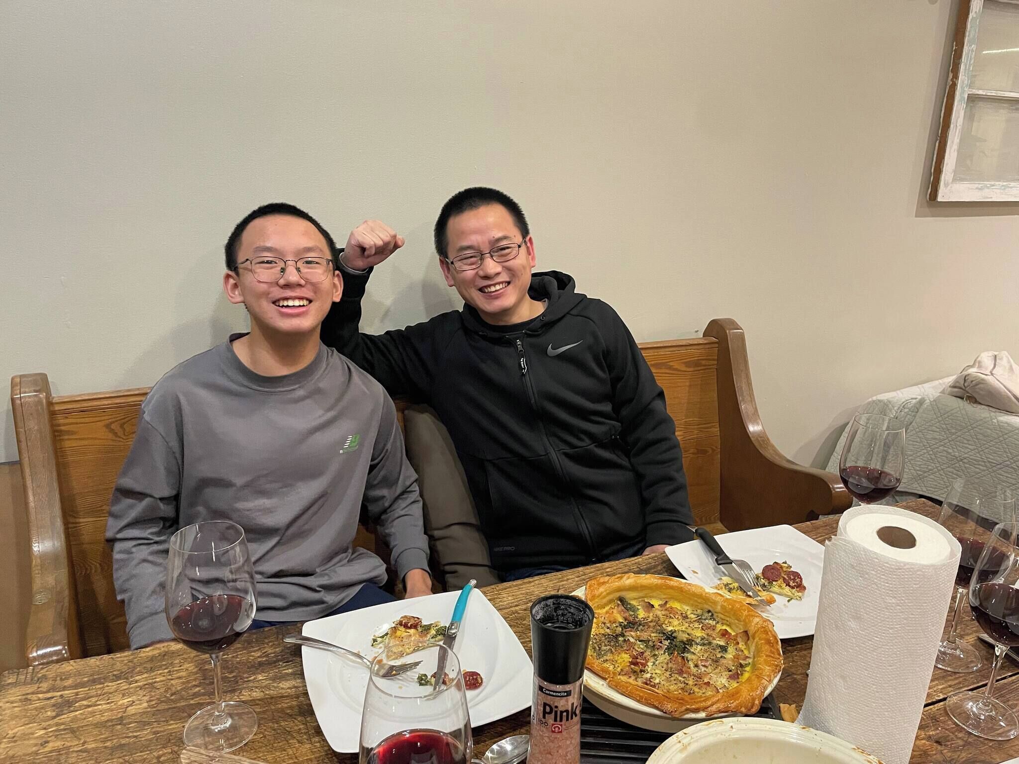 Two people seated at a wooden table with plates of food, wine glasses, and a paper towel roll.