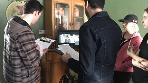 A group of people in a room with a wooden cabinet, a television, and some papers.
