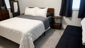 Un dormitorio con cama, mesitas de noche, un banco, un espejo y una ventana con cortinas.