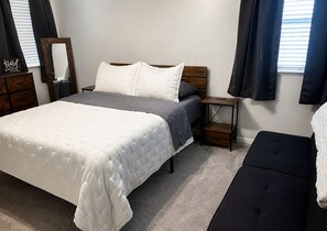 Ein Schlafzimmer mit Bett, Nachttischen, einer Bank, einem Spiegel und einem Fenster mit Vorhängen.