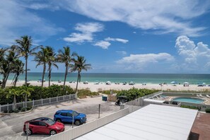 Blick auf einen Strand mit Palmen, einem Schwimmbecken und geparkten Autos.