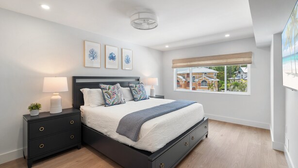 A bedroom with a bed, nightstands, a window with a view, and wall art. - Martinique South Unit 207 by SeaBreeze Vacation (Holmes Beach)