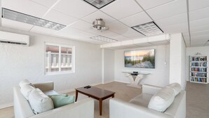 A modern living room with white furniture, a flat-screen TV, and a bookshelf. - Martinique South Unit 207 by SeaBreeze Vacation (Holmes Beach)