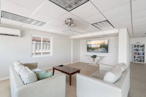 A modern living room with white furniture, a flat-screen TV, and a bookshelf. - Martinique South Unit 207 by SeaBreeze Vacation (Holmes Beach)