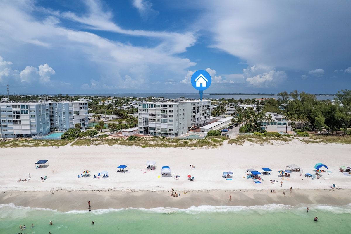 A beachfront hotel with multiple buildings, a sandy beach with umbrellas, and a clear view of the ocean.