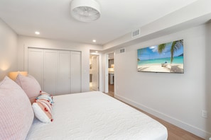 A bedroom with a bed, pillows, a TV on the wall, and a door leading to a bathroom. - Martinique South Unit 306 by SeaBreeze Vacation (Holmes Beach)
