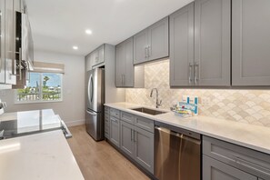 A modern kitchen with grey cabinets, stainless steel appliances, and a large window offering a view of palm trees. - Martinique South Unit 306 by SeaBreeze Vacation (Holmes Beach)