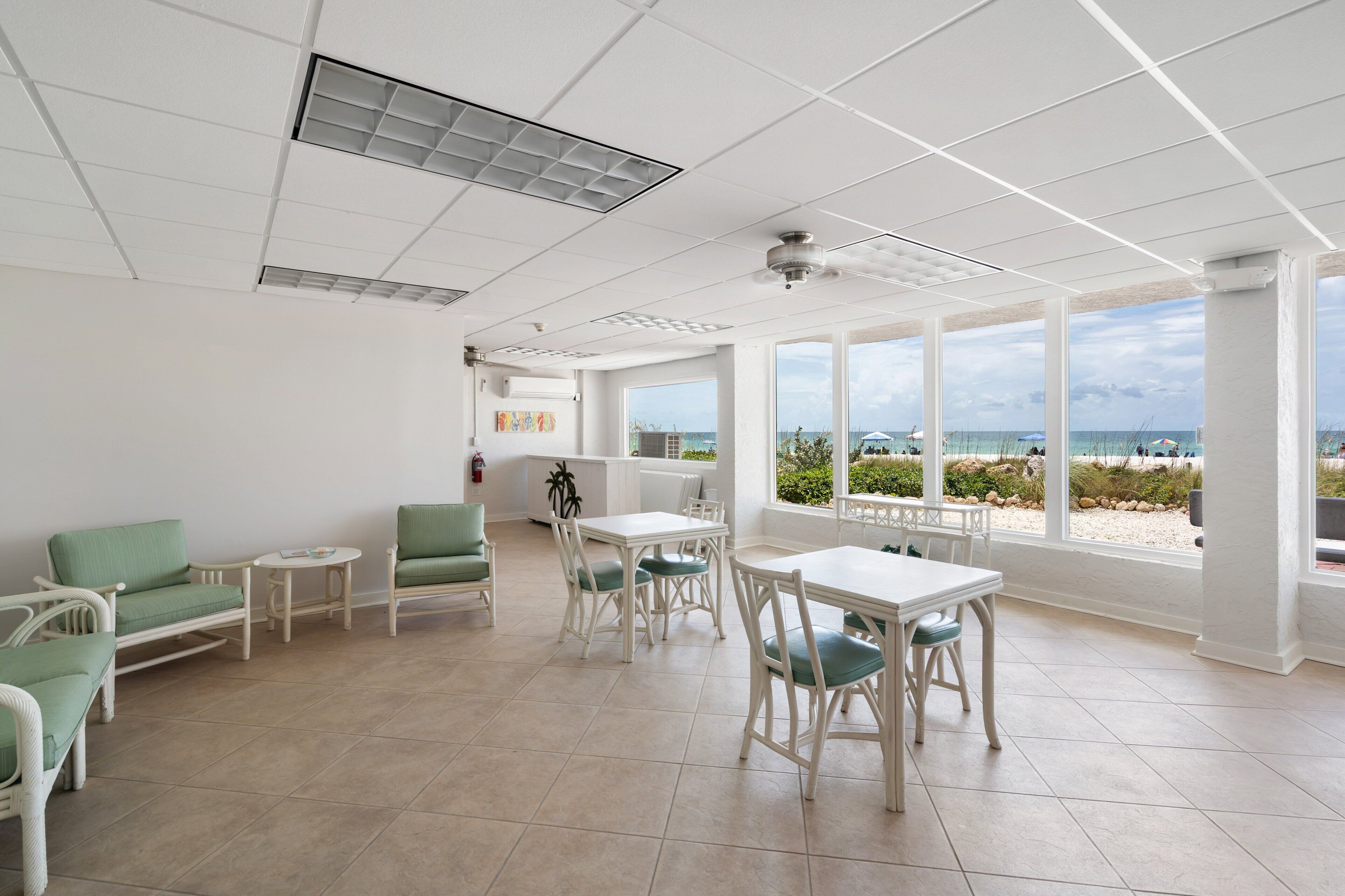 Ein geräumiger Raum mit großem Fenster und Blick zum Strand, weiße Decke mit Lüftungsgitter sowie ein weißer Tisch mit Stühlen.