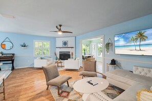 Un salon avec une cheminée, un ventilateur de plafond et un tableau sur le thème de la plage.