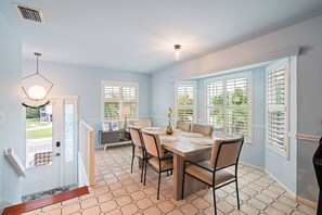 Un espace repas comprenant une table et des chaises, de grandes fenêtres équipées de volets et un luminaire.