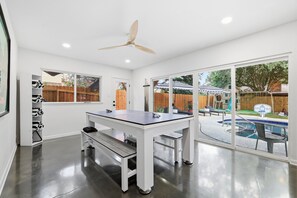 Ein Zimmer mit Tischtennisplatte, Deckenventilator und Blick auf einen Garten mit Schwimmbad.