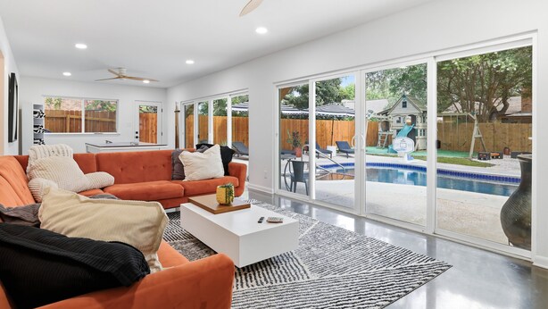 Ett modernt vardagsrum med en stor orange soffa, ett vitt soffbord och utsikt över en trädgård med pool.