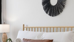 En säng med vita sängkläder, en sänggavel i trä, en flätad korg med handdukar och en svart, rund väggdekoration.