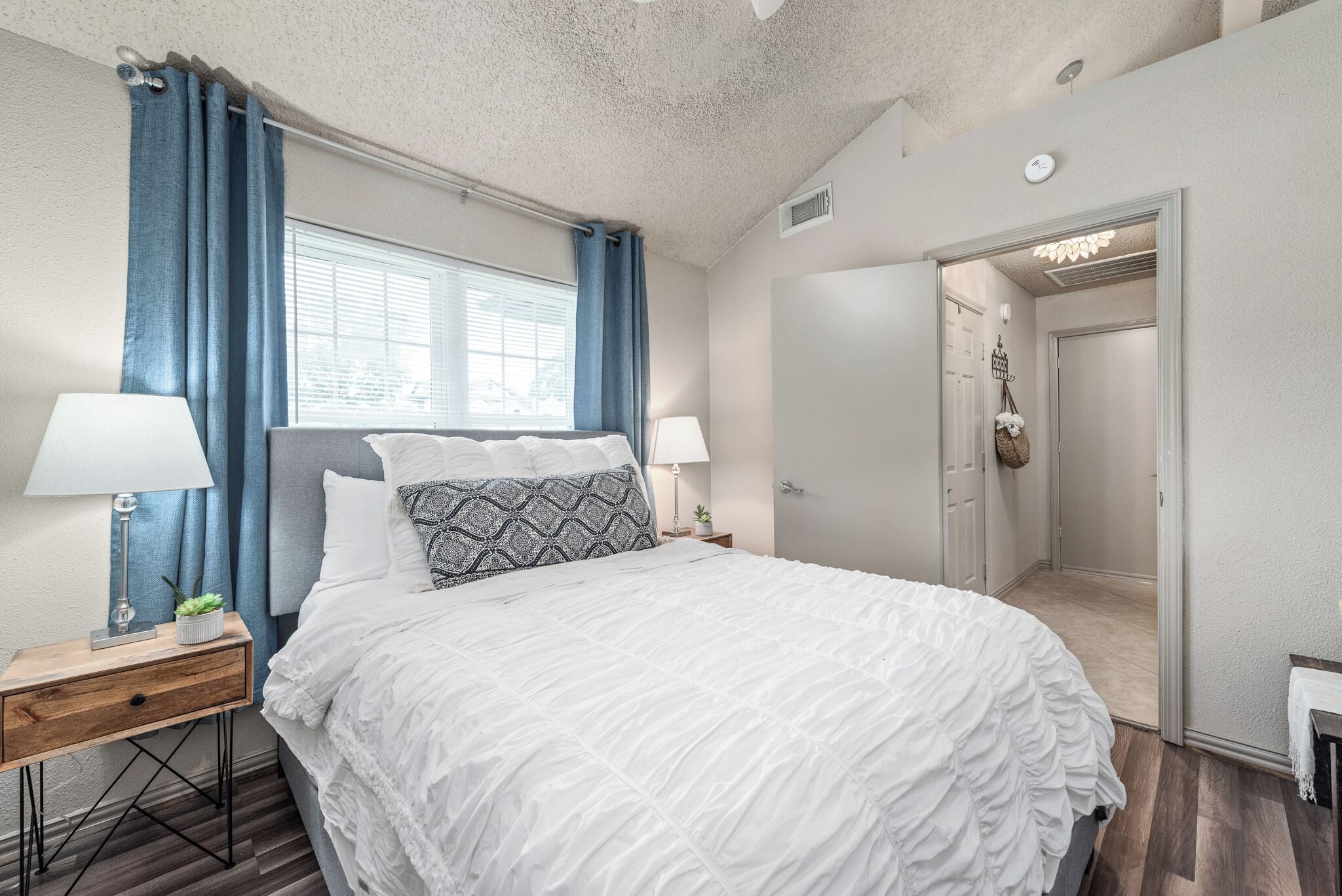 Een slaapkamer met een bed, twee nachtkastjes, een raam met gordijnen, een deur en een gang.