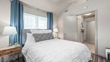 Een slaapkamer met een bed, twee nachtkastjes, een raam met gordijnen, een deur en een gang.
