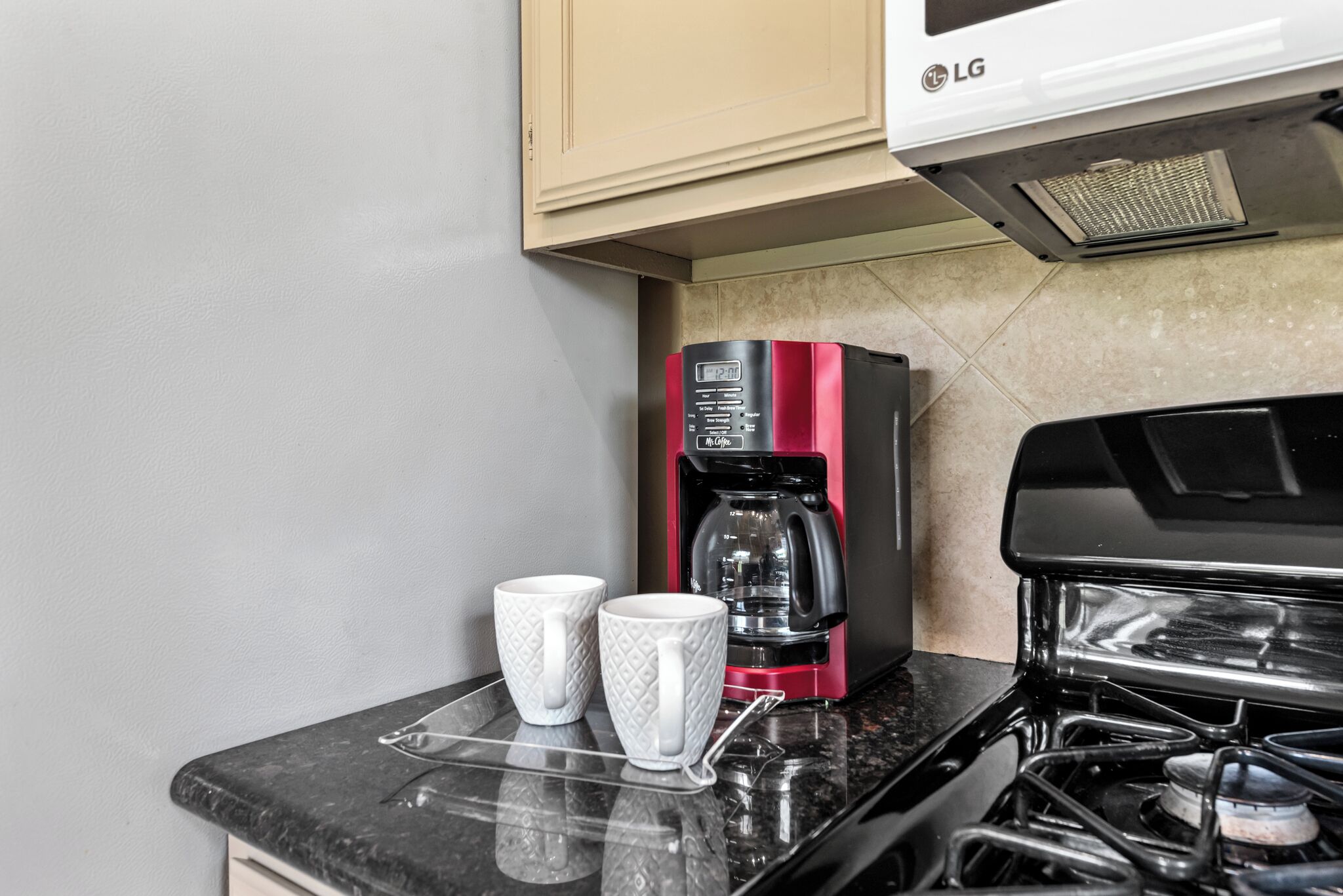 A kitchen with a gas stove, a coffee maker, and two white mugs on a black countertop.