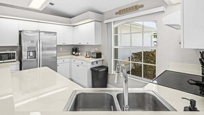 A modern kitchen with white cabinetry, stainless steel appliances, a black trash bin, and a window with a view. - Waterfront Gulf Access Retreat with Heated Pool, Dock & Outdoor Fun - Villa Snowscape - Roelens (Cape Coral)