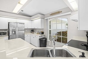 A modern kitchen with white cabinetry, stainless steel appliances, a black trash bin, and a window with a view. - Waterfront Gulf Access Retreat with Heated Pool, Dock & Outdoor Fun - Villa Snowscape - Roelens (Cape Coral)