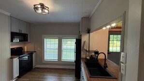 A kitchen with a microwave, oven, and sink. There are two windows with blinds, a ceiling light fixture, and a door leading to another room.