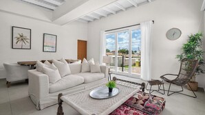 Un salon spacieux avec un canapé blanc, une table basse agrémentée d’une plante, un fauteuil en osier et une grande fenêtre avec des rideaux.