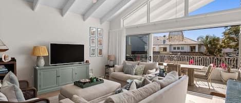 A spacious living room with a flat-screen TV, comfortable seating, and a balcony with a view of the neighborhood.