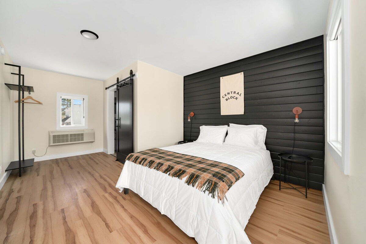 Quarto com cama, cabeceira preta, janela, luminária pendente e ar-condicionado fixado na parede.