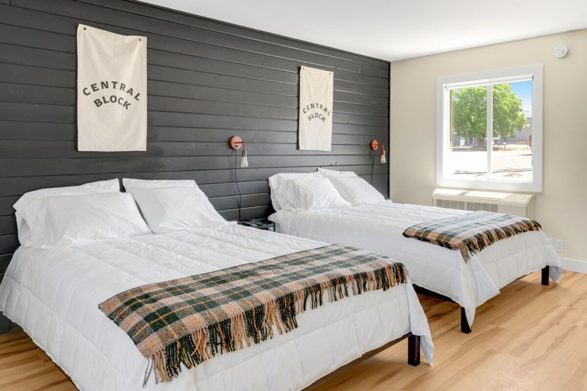 Quarto com duas camas, uma cabeceira de madeira escura, uma janela com vista e dois painéis decorativos na parede.