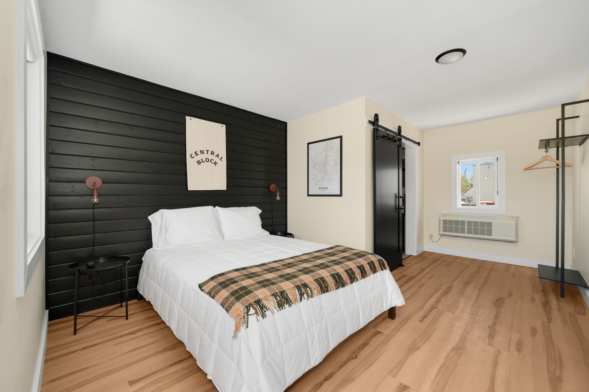 A bedroom with a bed, a nightstand, a wall-mounted light, a map, a black wardrobe, and a window.