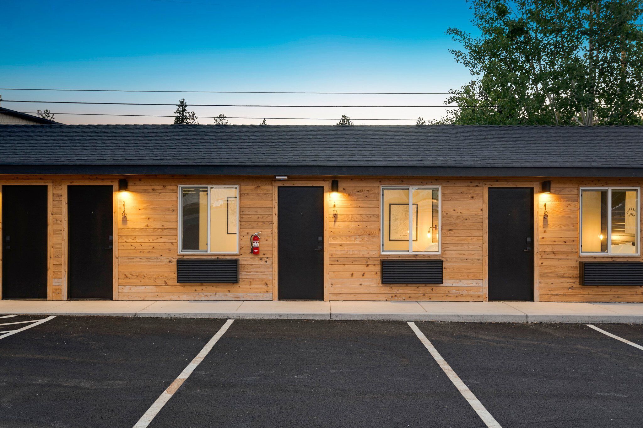 A row of log cabin-style rooms with individual windows and doors, a fire extinguisher mounted on the wall, and a parking lot in front.