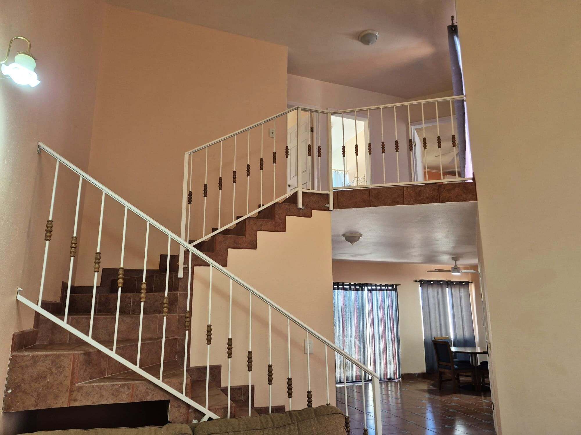 An interior perspective centered on a tiled staircase with white safety railings, leading up to a secondary loft space overlooking the lower dining area.