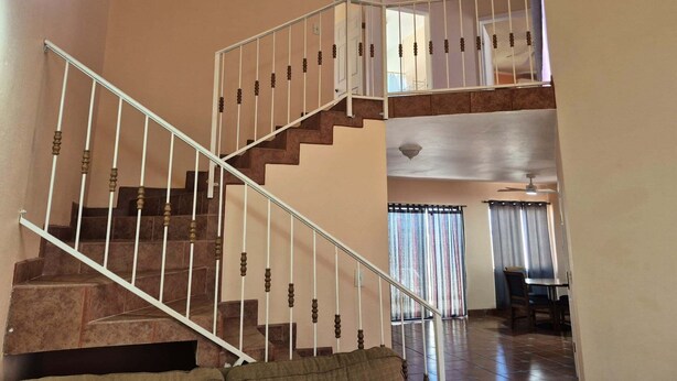 Uma escadaria com corrimão branco que leva a um segundo andar, uma área de refeições com mesa e cadeiras, e uma janela com cortinas.