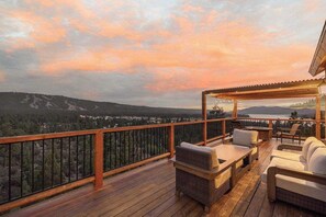 A wooden deck with outdoor furniture, a view of a lake, and a forested landscape. - Golden Oak Estate | 7BR Luxury. Wine Room. Views. (Big Bear Lake)