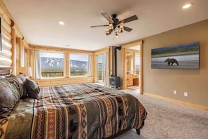Ein Schlafzimmer in einer Blockhütte mit einem großen Bett, einem Deckenventilator, einem Kamin und einem Fenster mit Blick auf die Berge.