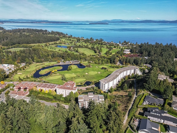 An aerial view of a golf course with a large building complex, surrounded by dense forest and a body of water.