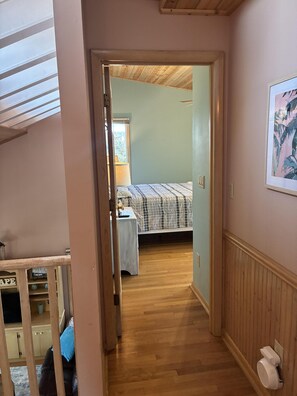 A narrow hallway leading to a bedroom with a bed, a window, and a wooden floor. - Welcome to Holden It Down!!!, your perfect coastal escape on the serene canals of Holden Beach. (Supply)