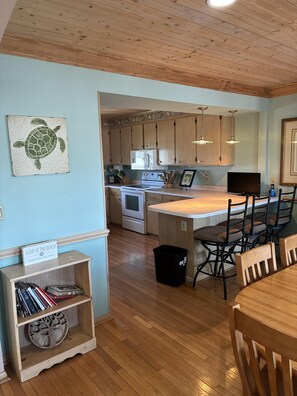 A kitchen with wooden cabinets, a microwave, a stove, and a dining area with chairs. - Welcome to Holden It Down!!!, your perfect coastal escape on the serene canals of Holden Beach. (Supply)