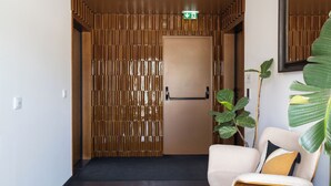 Um hall de hotel com divisória de madeira, balcão de recepção, uma cadeira com almofada e uma planta grande.