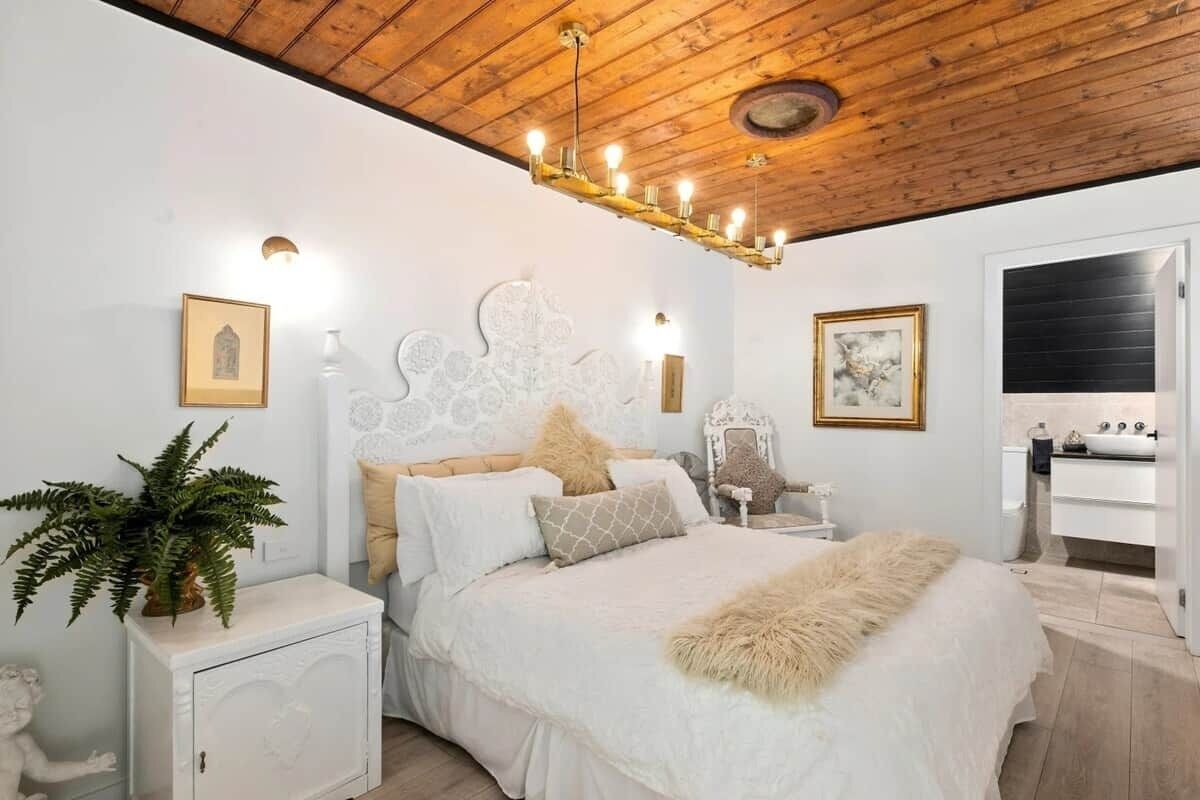 A bedroom with a wooden ceiling, a white bed with a fluffy blanket, a nightstand with a plant, and a white cabinet.