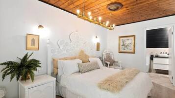 A bedroom with a wooden ceiling, a white bed with a fluffy blanket, a nightstand with a plant, and a white cabinet.