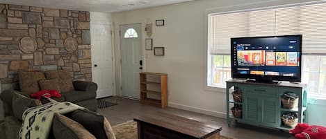 A living room with a stone wall, a sofa, a coffee table, a television on a stand, and a window with blinds.