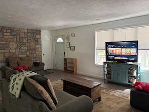 A living room with a stone wall, a sofa, a coffee table, a television on a stand, and a window with blinds.