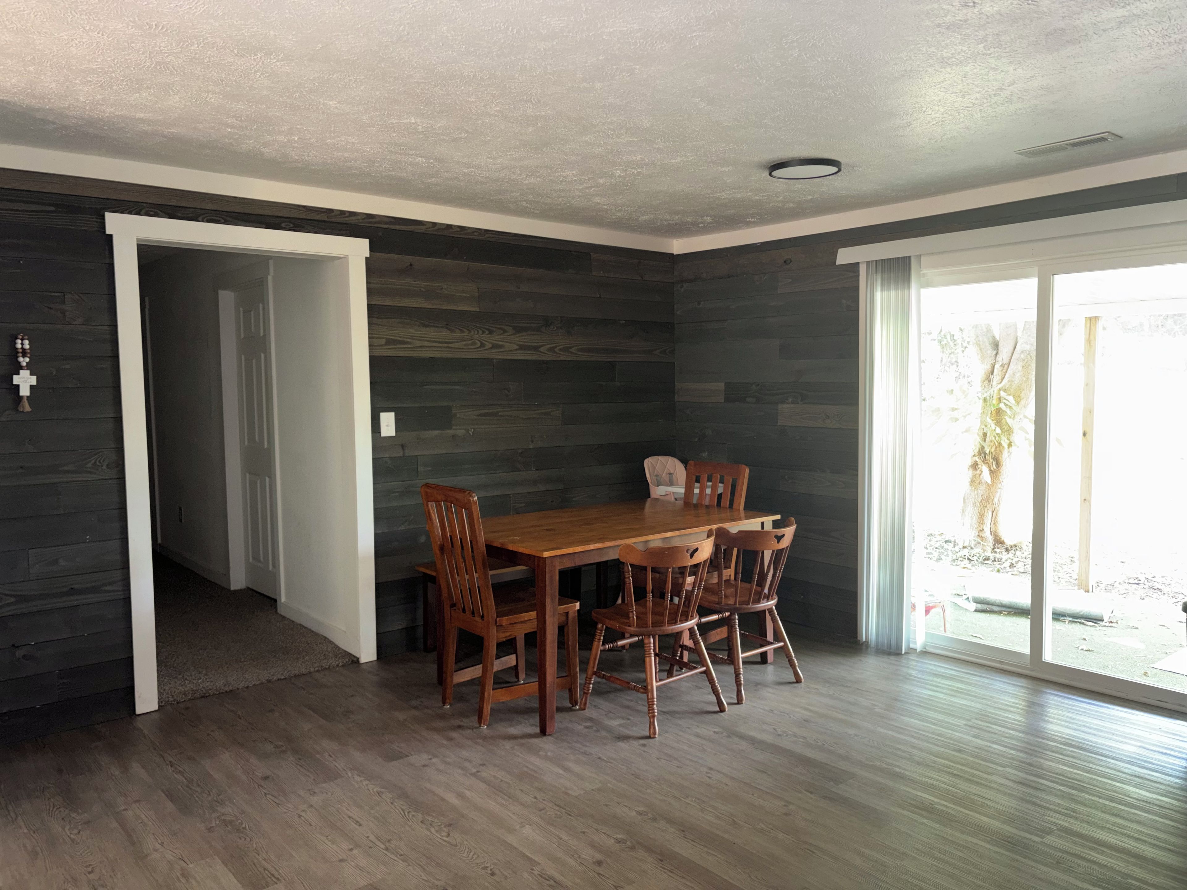 A dining area with a wooden table and chairs, a dark wood-paneled wall, and a sliding glass door leading to an outdoor area.