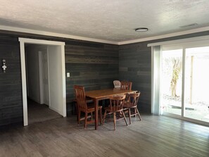 A dining area with a wooden table and chairs, a dark wood-paneled wall, and a sliding glass door leading to an outdoor area.