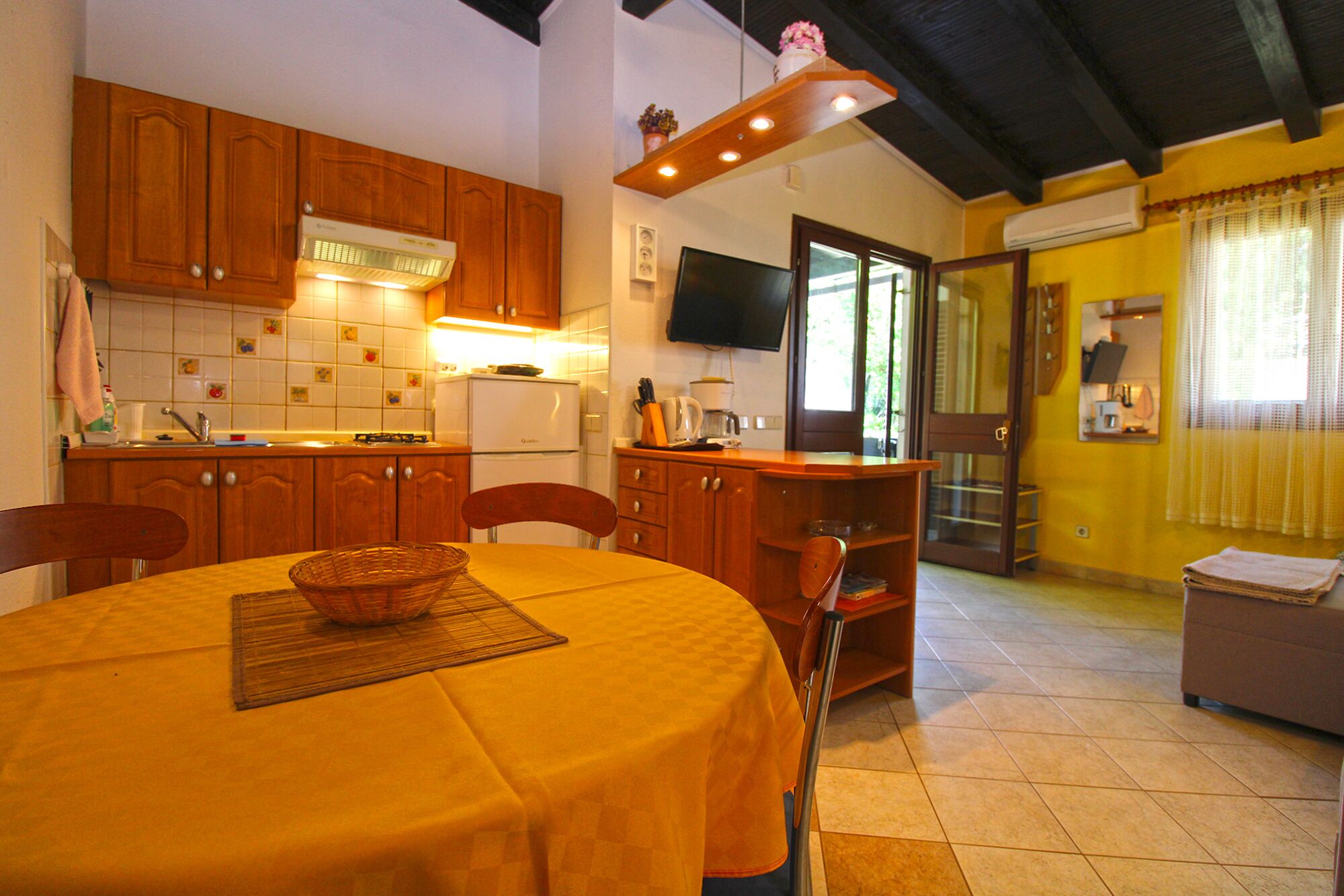 Una cocina con armarios de madera, una mesa de comedor con mantel amarillo y un televisor montado en la pared.