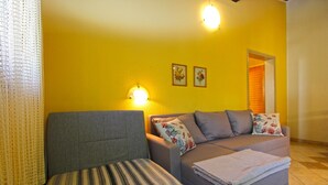 Una sala de estar con un sofá gris, un banquillo estampado y un sofá gris con cojines florales. Hay dos luces fijadas en la pared y dos cuadros enmarcados.