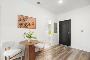Une pièce avec une table à manger en bois, deux chaises blanches, une plante en pot, une porte noire et une œuvre d’art accrochée au mur.