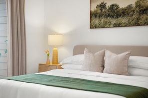 Ein ordentlich gemachtes Bett mit weißen Leinen, einer grünen Tagesdecke und zwei Kissen. Ein Nachttisch mit Lampe, einer Blumenvase und einer Kerze steht daneben. An der Wand hängt ein Landschaftsbild.